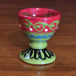 Può includere: Un portauovo in ceramica dipinto a mano con un design vibrante. La tazza presenta un interno e un bordo rossi, con fasce decorative verdi e blu. La base è verde con accenti neri e bianchi. Un pezzo decorativo.