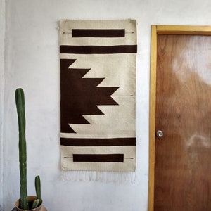 Puede incluir: Un tapiz de pared con estampado geométrico marrón y blanco con borde de flecos. El tapiz está colgado en una pared blanca.