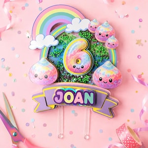 Puede incluir: Un adorno para tarta en capas que presenta un arcoíris, nubes blancas y cinco personajes sonrientes con estampado tie-dye. Un número seis central con acabado tie-dye se sitúa sobre una pancarta morada con el nombre JOAN en letras coloridas.
