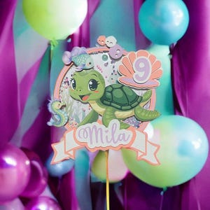 Könnte beinhalten: Ein Tortenaufsatz mit einer Cartoon-Meeresschildkröte, dem Namen "Mila" auf einem Banner und der Zahl 9. Das Design umfasst ein Seepferdchen und Fische. Der Hintergrund zeigt lila und türkisfarbene Vorhänge und bunte Luftballons.