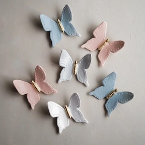 Op de afbeelding: Zes keramische vlinder wanddecoraties in verschillende pastelkleuren, waaronder lichtblauw, roze en wit. Elke vlinder heeft een gouden accent in het midden.