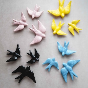 Pode incluir: Uma coleção de dez figuras de andorinha de cerâmica em várias cores, incluindo preto, rosa, azul e amarelo. As andorinhas estão dispostas em um padrão simétrico sobre um fundo cinza claro.