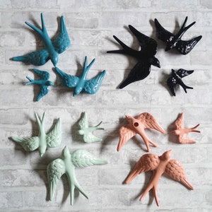 Puede incluir: Un conjunto de decoración de pared de golondrinas de cerámica en varios colores, incluyendo azul verdoso, negro, verde menta y melocotón. Las golondrinas están dispuestas en un patrón circular en una pared de ladrillo blanco.