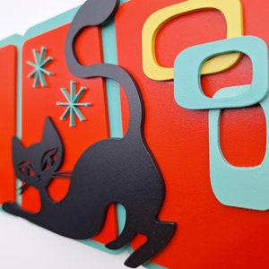 Puede incluir: Una silueta de gato negro con un fondo geom&eacute;trico de color verde azulado y rojo. El gato est&aacute; en una pose juguetona con la cola enroscada. El fondo presenta formas abstractas en verde azulado y amarillo.