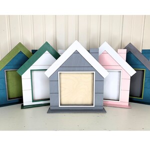 May include: A collection of house-shaped picture frames in various colors, including blue, green, gray, and pink. Each frame has a white roof and a rectangular opening for a photo. The frames are displayed on a white surface.