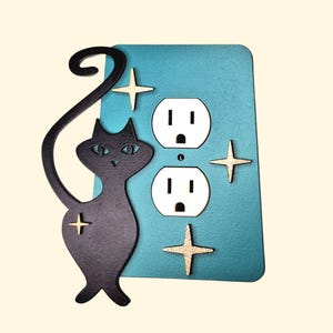 May include: A teal blue outlet cover with a black silhouette of a cat and gold stars. The outlet cover has two standard electrical outlets.
