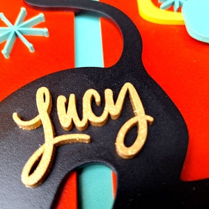 Puede incluir: Corte de madera negro y dorado de un gato con el nombre "Lucy" escrito en letras doradas con purpurina.