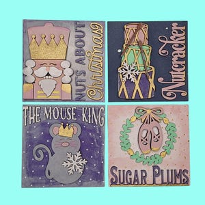 May include: Four square wooden signs with a Christmas theme. The signs are painted in pink, purple, green, and blue with gold glitter accents. The signs feature the text "Nuts About Christmas", "The Mouse King", "Sugar Plums", and "Nutcracker".