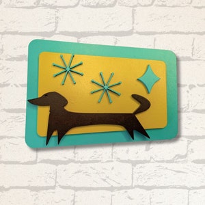 May include: A mid-century modern-style wall art piece featuring a brown dachshund silhouette on a yellow background, accented with turquoise starbursts and a diamond shape. The artwork is set against a turquoise border.