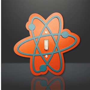 May include: Orange and teal light switch cover with an atomic symbol design. The switch is white.