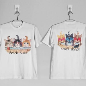 May include: White t-shirts with a colorful cat graphic. The front of the shirt features three cats with the text "Beach Bums". The back of the shirt shows the cats' backsides. The cats are in shades of orange, black, and blue.