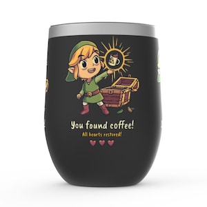 May include: A black and green stainless steel tumbler with a cartoon image of Link from the Legend of Zelda video game series. Link is holding a cup of coffee and a treasure chest is open behind him. The text "You found coffee! All hearts restored!" is printed on the tumbler.