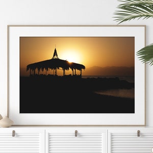 May include: A framed photograph of a silhouette of a thatched roof hut on a beach at sunset. The sun is setting behind the hut, casting a warm glow on the scene.