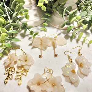 May include: A collection of handmade earrings featuring floral and leaf designs. The earrings are in shades of white and gold, with a shimmering, opalescent finish. Some earrings are hoop style, others dangle.