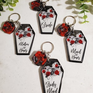 May include: Five coffin-shaped keychains with a red rose charm. Each keychain has a different title: Bride, Matron of Honor, and Bridesmaid. The coffin design features red roses and clockwork elements on a white background.