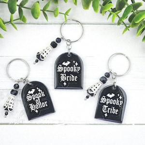 May include: Three black keychains with tombstone-shaped pendants. Each keychain has a silver ring, a decorative charm, and a pendant with white text: "Spooky Bride", "Spook of Horror", and "Spooky Tribe".