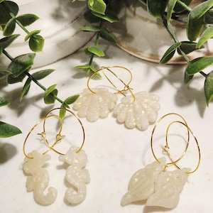 May include: Three pairs of earrings with gold hoops. One pair features seahorse charms, another has seashell charms, and the third has flower-shaped charms. All charms are white with glitter accents. Green leafy vines are in the background.