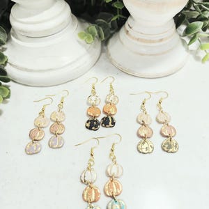 May include: A collection of dangle earrings featuring a pumpkin design. The earrings are in various colors, including white, orange, pink, black, and blue, with gold accents. The earrings are displayed on a white surface.