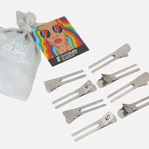 Puede incluir: Un juego de ocho pinzas para el pelo plateadas en una bolsa de cordón blanco con el texto "Shan Clips" impreso en ella. La bolsa está al lado de una tarjeta colorida con un dibujo de una mujer con el pelo arcoíris que lleva gafas de sol. La tarjeta también tiene el texto "Shan Clips" impreso en ella.