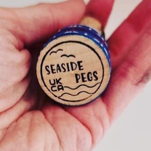 Könnte beinhalten: Ein hölzerner Flaschenverschluss mit blau-weiß gestreifter Oberseite. Der Verschluss trägt die Aufschrift "SEASIDE PEGS" und "UK CA" sowie eine Welle und drei Möwen.