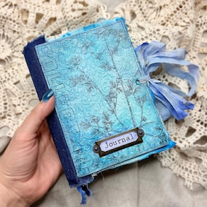 May include: A handmade journal with a textured, turquoise cover featuring a floral design and the word "Journal" on a metal label. The journal is bound with a dark blue spine and tied with a light blue ribbon.