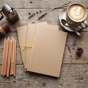Op de afbeelding: Een stilleven met briefpapier en een koffiepauze. Kraftpapieren notitieboekjes vastgebonden met geel touw, potloden, een puntenslijper en een kop koffie met latte art zijn gerangschikt op een verweerd houten oppervlak.