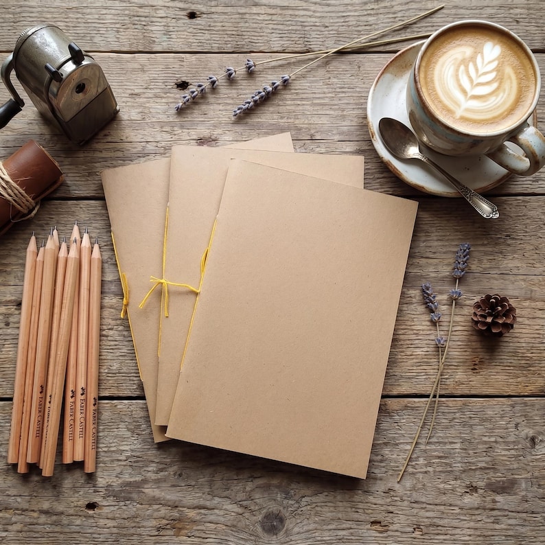Three stitched kraft notebooks sit on a wooden table beside coffee, pencils, and lavender. Perfect as a Weekly Planner, recipe book, or cute Undated Planner.