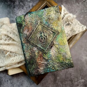 May include: A textured journal with a bronze dragon emblem. The cover features a mix of green, brown, and gold hues, with a decorative metal frame around the dragon. The journal is placed on a wooden frame with lace.