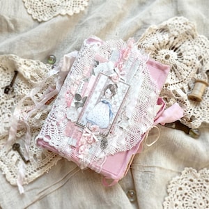 Pode incluir: Um diário rosa com uma capa decorativa com renda branca, fitas rosa e detalhes florais. Uma etiqueta com uma menina de desenho animado em um vestido está anexada. O diário é cercado por toalhas de renda e chaves vintage.
