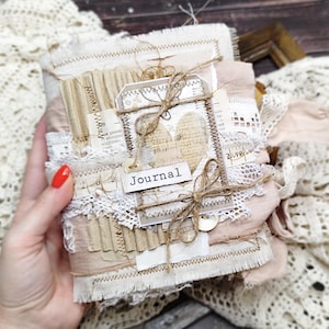 Könnte beinhalten: Ein handgefertigtes Journal mit einem Stoffeinband in Beige- und Cremetönen, verziert mit geschichtetem Papier, Spitzenbesatz, Wellpappe und Bindfaden. Ein kleines rechteckiges Schild mit dem Wort Journal ist mit einem Knopf befestigt.