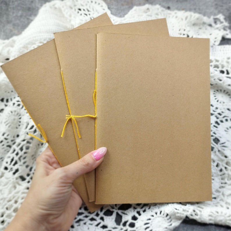 A hand holds three rustic kraft notebooks stitched with bright yellow thread. This versatile blank journal set is perfect for a travelers notebook or Craft junk journal.