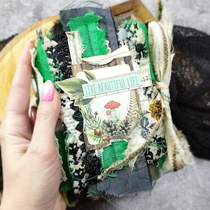 May include: A handmade journal with a green, black, and white fabric cover. The cover is decorated with a variety of embellishments, including a small tag with the text "Live Beautiful Life".