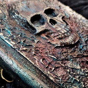 May include: A rectangular book cover featuring a raised, three-dimensional skull relief. The surface has a heavily distressed, weathered texture with a dark color palette of deep brown, bronze, and oxidized teal green highlights throughout.