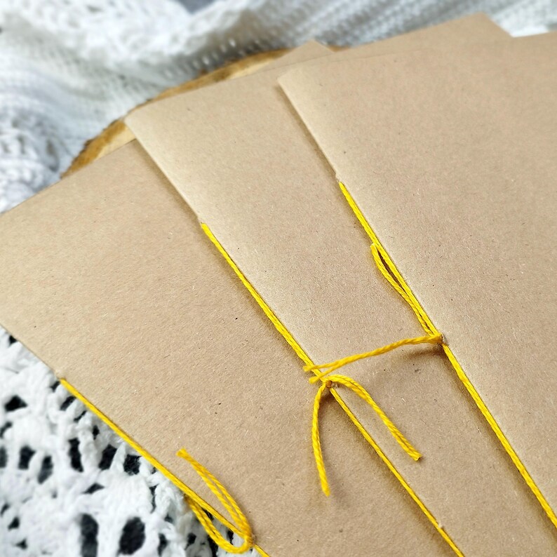 Close-up of three kraft covers with yellow thread binding, resting on white lace. These handmade books make excellent Journal refill paper for resurrected journals.