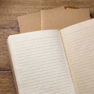 An overhead shot of an open rustic notebook on a textured wooden surface. The cream pages of the notebook are clearly lined for writing. In the background, on a small wooden cutout, lie two closed notebooks with brown kraft covers and a visible yellow thread spine.