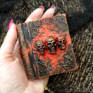 Puede incluir: Un pequeño libro texturizado con una apariencia oscura y envejecida. La portada está adornada con tres calaveras de color bronce y una superposición roja texturizada. Los bordes del libro tienen un acabado dorado, añadiendo un toque de elegancia al diseño gótico.