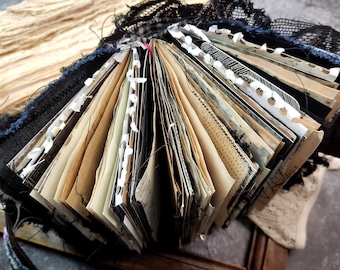 May include: A vintage-style journal with a black fabric cover and lace trim. The pages are filled with aged paper and have been decorated with white paper strips and stitching. The journal is open to reveal the pages.