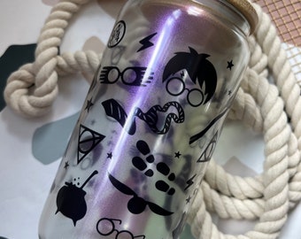 May include: A clear glass water bottle with a black bamboo lid and a straw. The bottle has a holographic iridescent finish and features black and white Harry Potter themed graphics, including glasses, a lightning bolt, a snake, a cauldron, and the Hogwarts crest.