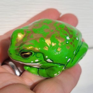 May include: A small green frog figurine with gold and white accents. The frog is sitting on a hand.