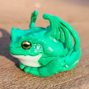 May include: A handcrafted, green frog figurine with dragon-like wings and horns. The frog has golden eyes and a white underbelly. The figurine is made of a glossy material and sits on a wooden surface.