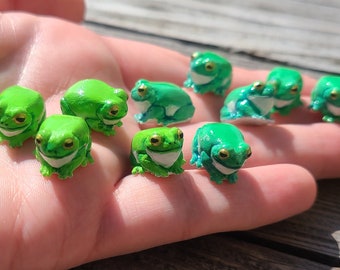 Puede incluir: Una colección de pequeñas figuritas de ranas verdes con vientres blancos. Las ranas están dispuestas en una mano, algunas mirando hacia adelante y otras de lado. Las ranas están pintadas con detalles realistas, incluidos ojos, patas y dedos de los pies.