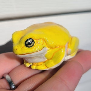 May include: A small, yellow frog figurine with black eyes. The frog is sitting on a hand.