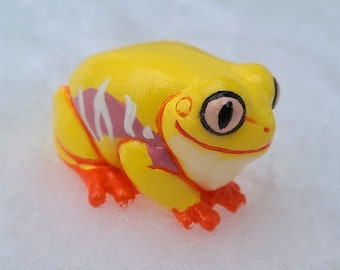 Könnte beinhalten: Eine kleine gelbe Froschfigur mit rosa und weißen Markierungen auf dem Rücken. Der Frosch hat große schwarze Augen mit rosa Pupillen und ein breites Lächeln.