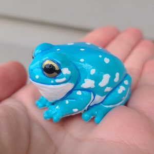 May include: A small, blue frog figurine with white spots. The frog has yellow eyes and a glossy finish.