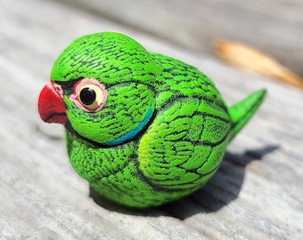 May include: A small green ceramic bird figurine with a red beak and black eyes. The bird has a detailed design of green and black feathers.
