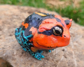 May include: A hand-painted ceramic frog figurine with a black, orange, and blue color scheme. The frog has a textured surface and a realistic appearance.