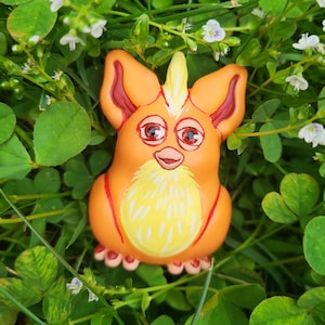 Könnte beinhalten: Orangefarbenes und gelbes flauschiges Spielzeug mit einem spitzigen orangefarbenen Irokesenschnitt. Das Spielzeug hat rote Augen und einen rosa Mund. Das Spielzeug sitzt auf einem Bett aus grünem Klee.