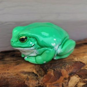 May include: A green ceramic frog figurine with white spots and gold eyes. The frog is sitting on a brown wooden surface.