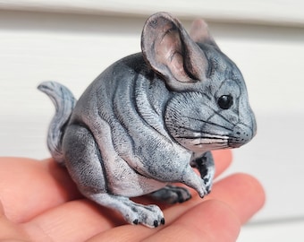 May include: A gray and white ceramic figurine of a chinchilla. The figurine is sitting on a hand and has a fluffy tail.