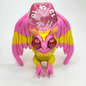 May include: A whimsical, hand-painted figurine of a fantasy creature with pink and yellow wings, a yellow body, and large red eyes. A pink, transparent, twenty-sided die rests on its head. The figurine is designed for tabletop gaming.
